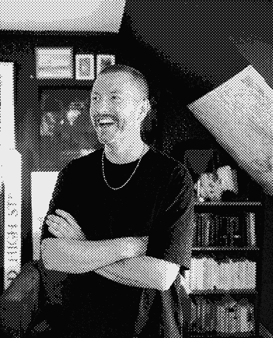 Stu by the studio bookshelf, arms crossed, caught mid-laugh